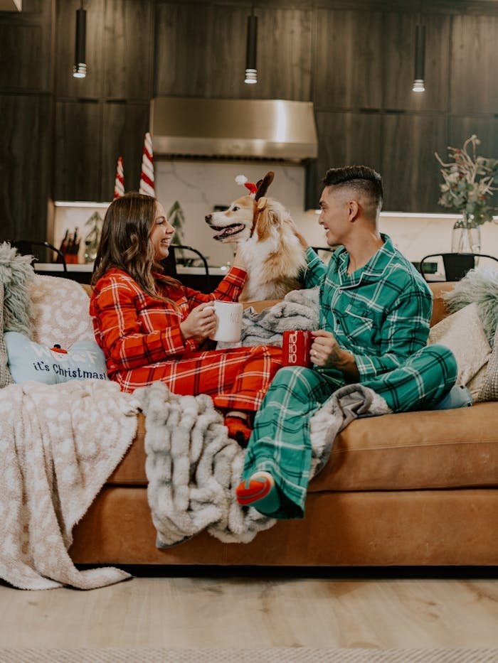 Cozy couple in plaid onesies sharing coffee with their dog on Christmas morning.
